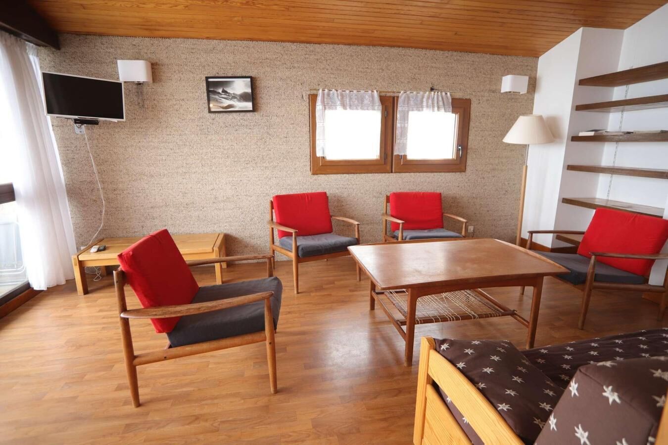 A cozy living room with wooden furniture, a flat-screen TV, and two windows with curtains.