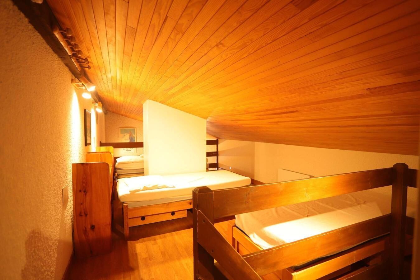 A small, cozy attic bedroom with wooden beams, two bunk beds, a wooden wardrobe, and a small table with a lamp.