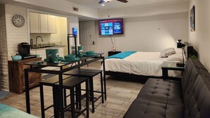 Een ruimtelijk ingerichte woonruimte met een kitchenette, een bed met een blauwe deken, een bank en een plafondventilator.