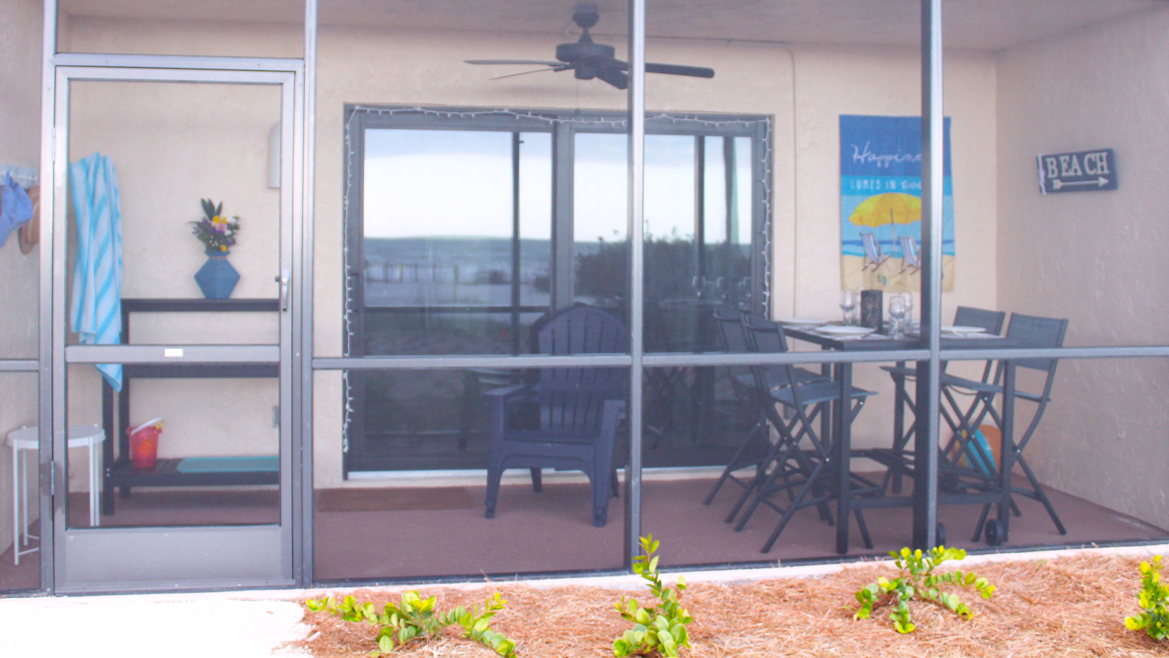 A covered outdoor dining area with a table and chairs, a ceiling fan, and a view of the beach through a glass door.