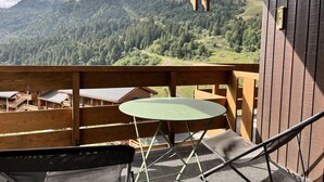A wooden balcony with a green table and a chair, overlooking a mountainous landscape with buildings in the background. - 5m from ski slopes, floor -2, view mountain, balcony, 34m², Méribel-Mottaret (Méribel - Les Allues)
