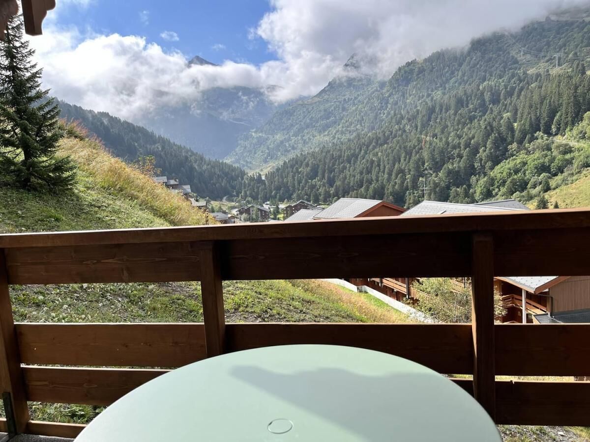 Una terraza de madera con una mesa y sillas, con vistas a un paisaje montañoso que incluye casas y un valle.