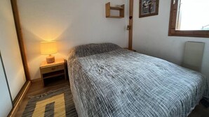 A bedroom with a bed, a nightstand with a lamp, a wooden shelf, and a window. - 5m from ski slopes, floor -2, view mountain, balcony, 34m², Méribel-Mottaret (Méribel - Les Allues)