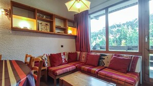 Un salon chaleureux avec un canapé en bois, une table basse en bois et une table à manger avec des chaises. Des étagères sont disposées au-dessus du canapé, où l’on peut voir des objets décoratifs, et une fenêtre est agrémentée de rideaux.