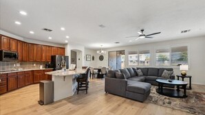 A spacious living area with a gray sectional sofa, a coffee table, and a dining area with wooden cabinets and a microwave. - Spacious 5-bedroom house with Heated Pool, hot tub and fire pit. (Goodyear)
