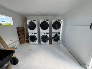 Un cuarto de lavandería con tres lavadoras de carga frontal apiladas, una puerta de madera y una ventana que da a vistas de vegetación.