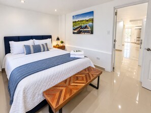 A modern hotel room with a large bed, a wooden bench, a nightstand with a lamp, and a painting on the wall. - Breathtaking Beachfront Gem in Luquillo, WAVES  at Your Door.Centerally Located to Rainforest, BioBay, Ferry Trips, Airport, Horses, Golf, KIOSK. Your Charming Home is Fenced and Gated for Privacy, Right on the BEACH and a Blast of Family/Friend Fun. (Playa Fortuna)