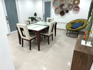Un comedor con una mesa de vidrio, cuatro sillas, una silla verde con cojín azul y una pared decorada con cestas tejidas.