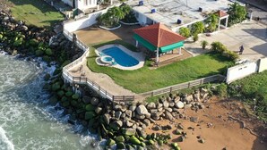 An aerial view of a coastal property with a swimming pool, a covered patio, and a beach area with rocks and a sandy shore. - Breathtaking Beachfront Gem in Luquillo – Walk to Kiosks & Hidden Bars (Playa Fortuna)