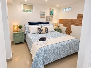 Un dormitorio con cama, mesitas de noche, lámpara, planta y pared de ladrillo.