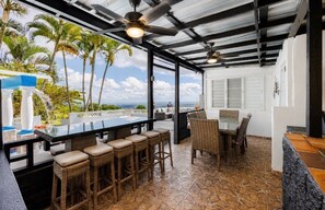 Un espace bar extérieur avec vue sur les palmiers et l’océan, agrémenté de meubles en osier et d’une table à plateau en verre.