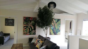 Una sala de estar moderna con un sofá gris, una mesa de centro y una planta grande. Hay pinturas abstractas en las paredes y una lámpara en el techo.