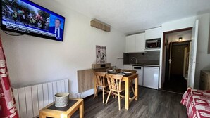 Un espai vital compacte amb zona de menjador, petita cuina i televisió que mostra un informatiu.
