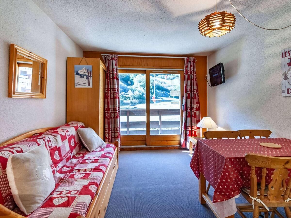 Une pièce chaleureuse avec un canapé en bois, une table à manger recouverte d’une nappe rouge et une grande fenêtre agrémentée de rideaux.
