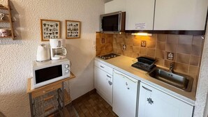 A compact kitchen with a microwave, coffee maker, and a stove. There are framed botanical prints on the wall and a small shelf with kitchen items.