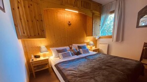 Un dormitorio acogedor con cabecera de madera, una cama grande con manta oscura, dos mesitas de noche con lámparas y una ventana con cortinas.