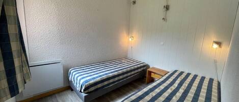 Een kleine, compacte kamer met twee eenpersoonsbedden, een houten nachtkastje en een wandradiator.
