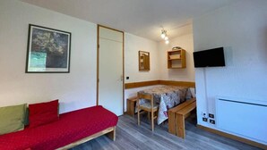 Une petite pièce meublée, comprenant un canapé, une table à manger avec des chaises, un téléviseur fixé au mur et une porte donnant sur une autre pièce.