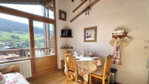 Um quarto aconchegante com uma mesa de jantar de madeira, cadeiras, uma cama com colcha xadrez, uma janela com vista para o campo, um relógio de parede, um quadro emoldurado e uma estante com uma televisão e outros objetos.