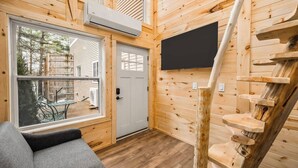 Ein gemütliches Blockhäuschen im Inneren, mit Holzvertäfelung, einer Treppe, einem an der Wand montierten Fernseher und Blick ins Freie.