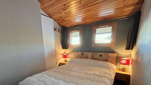 Un dormitorio con techo de madera, una cama blanca, dos mesitas de noche con lámparas rojas y dos ventanas con cortinas.