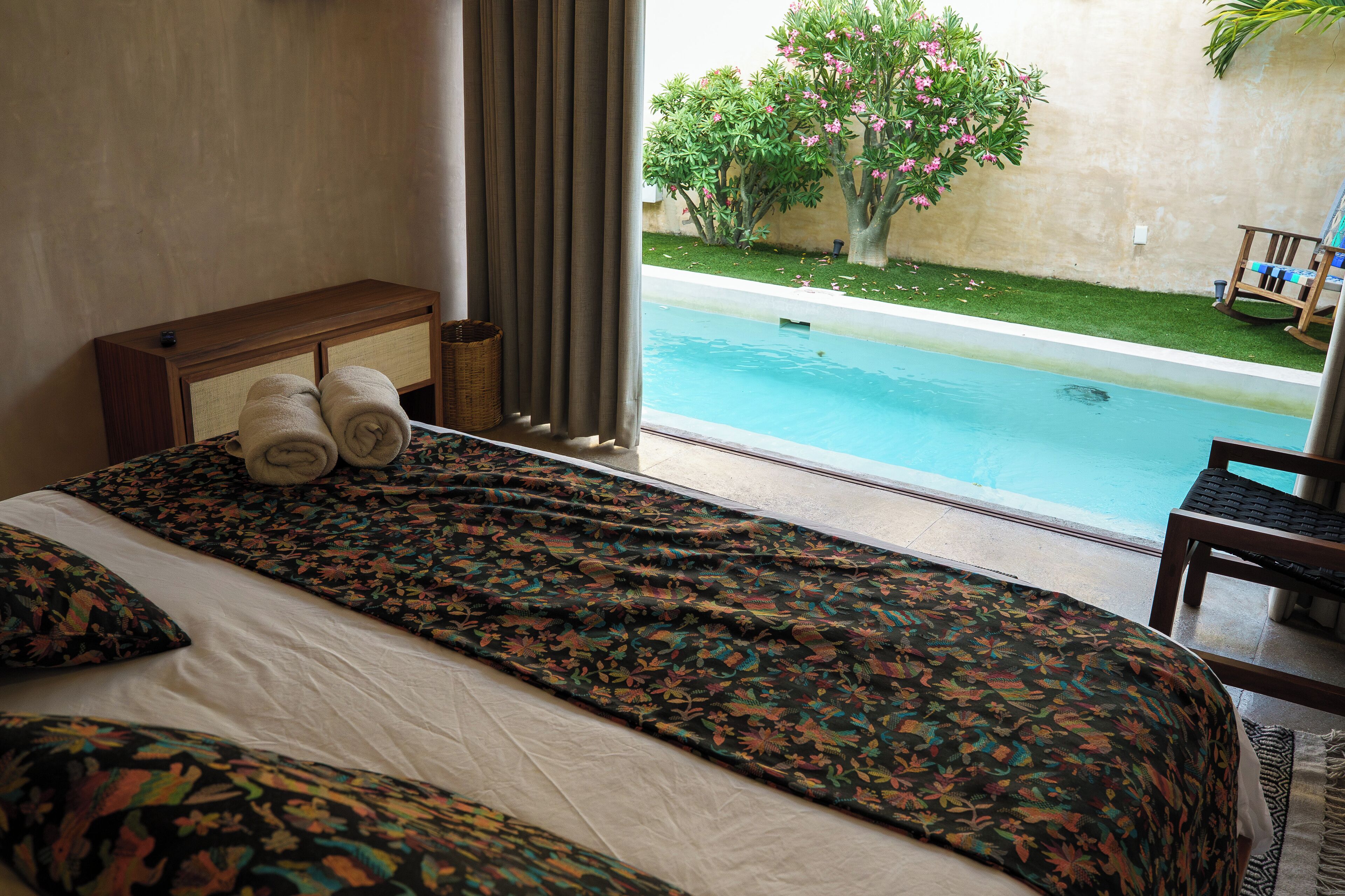 A hotel room with a bed, a window with curtains, a view of a swimming pool, and a chair outside.