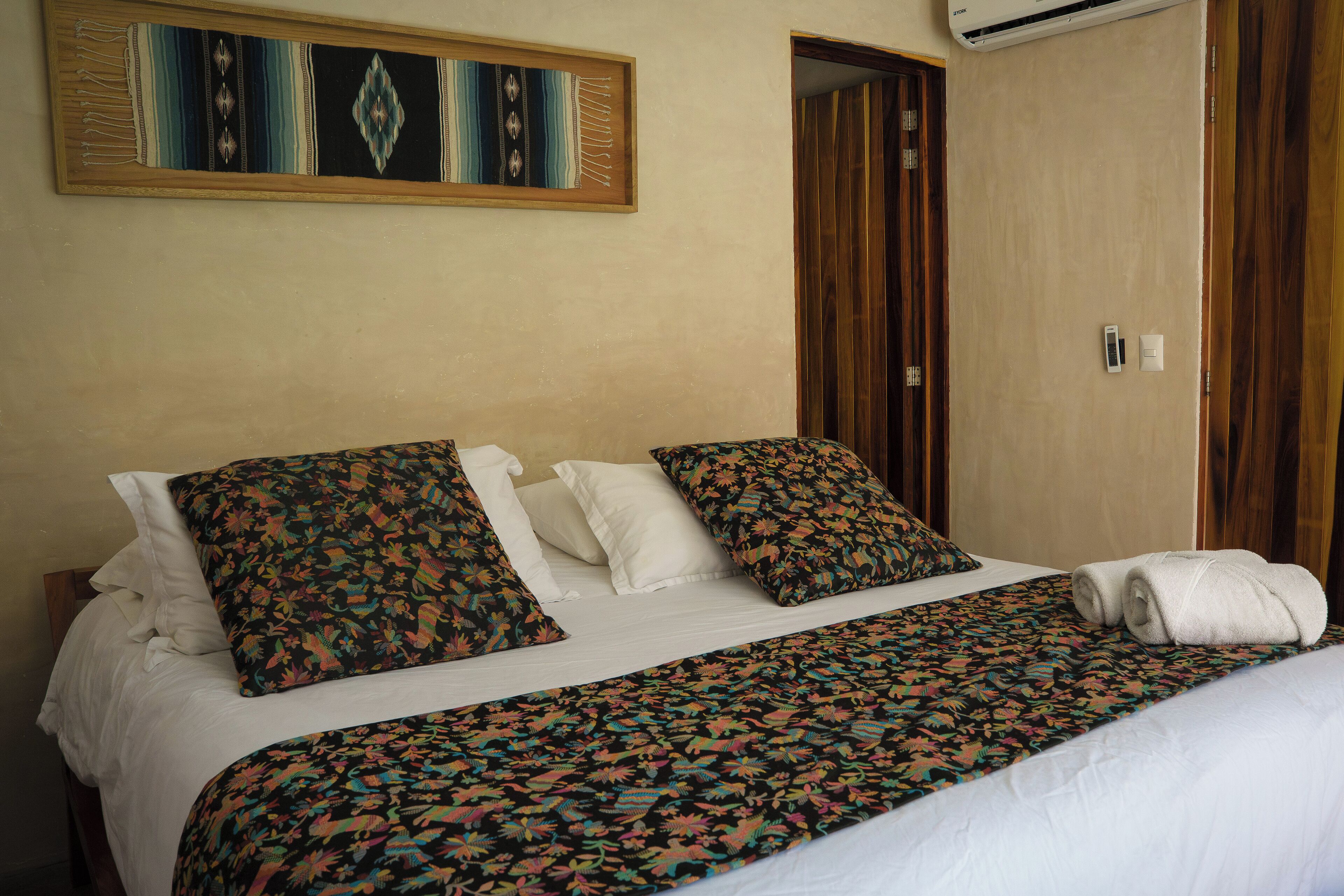 A neatly made bed with a floral-patterned bedspread and pillows, a framed tapestry on the wall, and a wooden door with a light switch and thermostat.