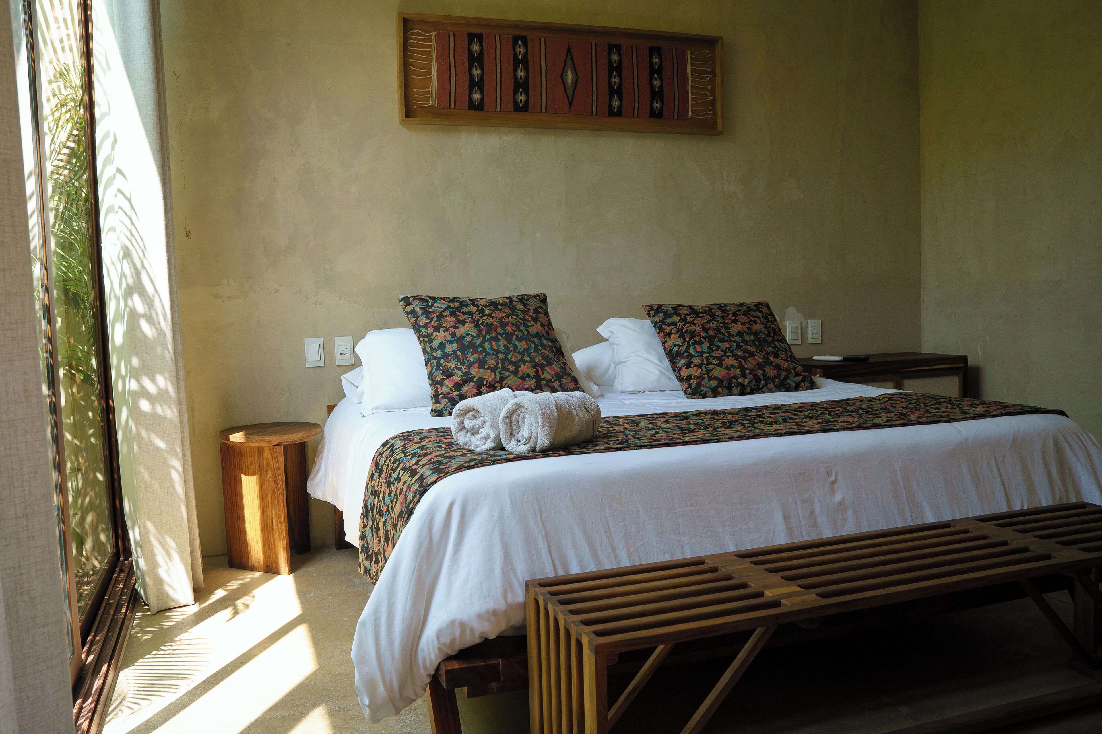 A neatly made bed with white linens and patterned pillows, a wooden bench, and a small round table beside the bed.
