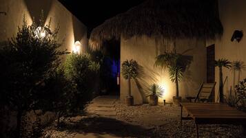 A nighttime view of a courtyard with a thatched-roof building, potted plants, a wooden bench, and a stone pathway.