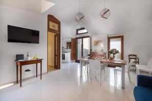A spacious living area with a flat-screen TV, a dining table with chairs, a kitchen area, and a large window.