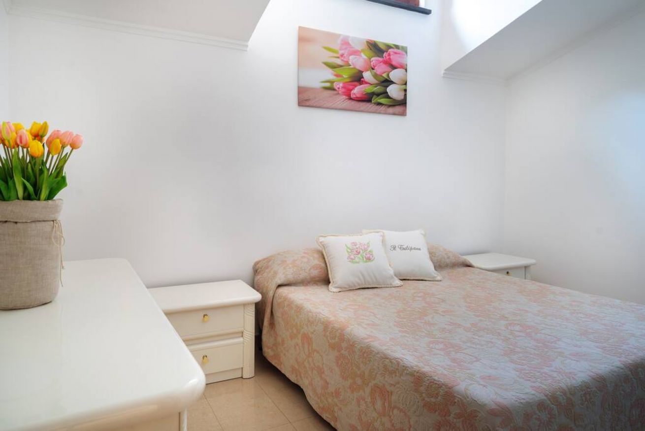 Una camera da letto con un letto, comodini, un tavolino da notte, un quadro di fiori appeso al muro e una finestra.