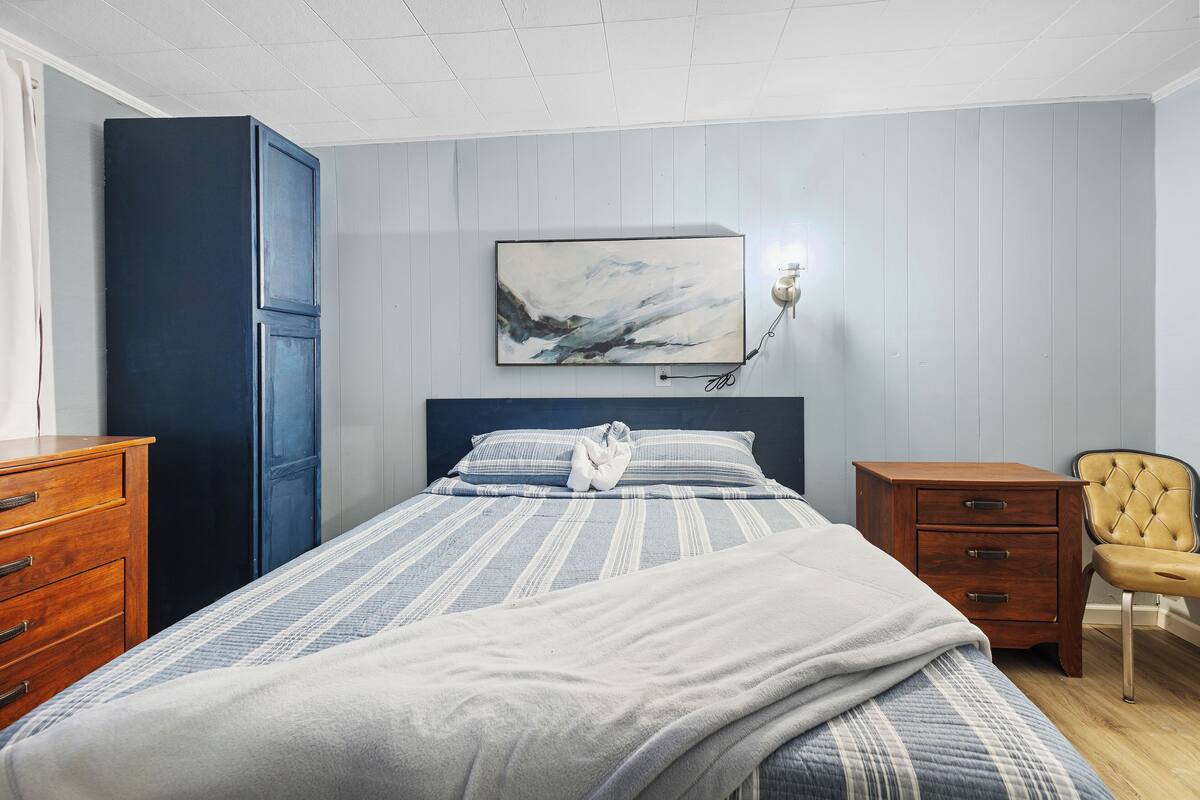 A bedroom with a bed, a nightstand, a chair, a wardrobe, and a wall-mounted artwork.