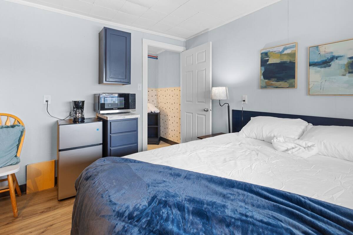 A hotel room with a bed, a desk with a microwave, a refrigerator, a chair, and a door leading to a bathroom.