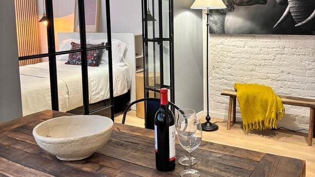 Een moderne woonkamer met een houten eettafel, twee zwarte hanglampen, een wijnfles en een glas. Een grote muurschildering van een olifant versiert de wand, en een gele plaid ligt over een bank.