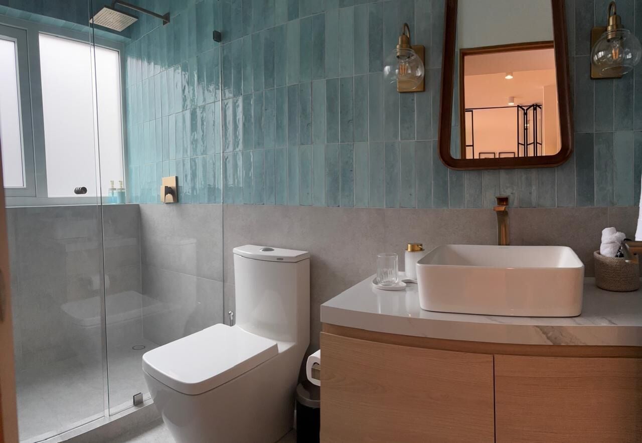 Un baño moderno con mampara de vidrio, inodoro blanco y un lavamanos rectangular grande sobre un mueble de madera.