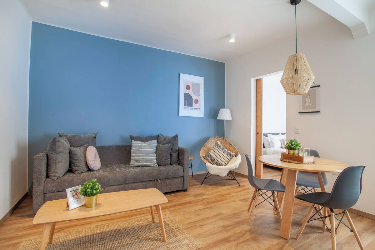 A modern living room with a blue accent wall, a grey sofa, a wooden coffee table, and a dining area with a round table and chairs.