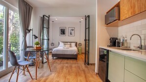 Een moderne hotelkamer met een houten vloer, een bed met witte lakens, een eettafel met stoelen, een keuken met een magnetron en uitzicht op groen door grote ramen.