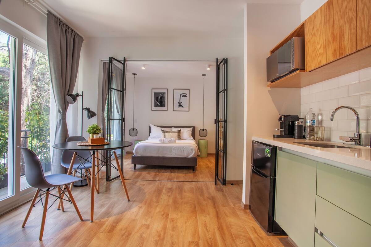 A modern hotel room with a wooden floor, a bed with white linens, a dining table with chairs, a kitchen area with a microwave, and a view of greenery through large windows.