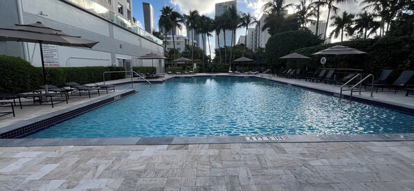 A swimming pool with lounge chairs and umbrellas, surrounded by palm trees and high-rise buildings.