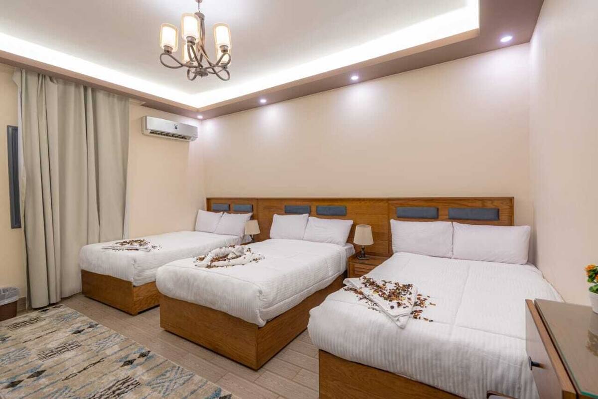A hotel room with two single beds, a larger double bed, a wooden headboard, a chandelier, an air conditioning unit, and a small table with a vase.