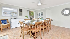 Uma sala de jantar com uma mesa posta para seis pessoas, rodeada por cadeiras de vime, um ventilador de teto e vista para o exterior através de uma janela.