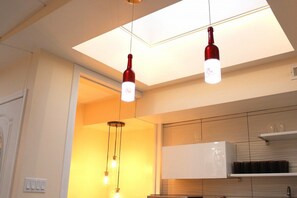 Une cuisine avec des luminaires suspendus au plafond, un réfrigérateur blanc et une étagère où sont posées des tasses.