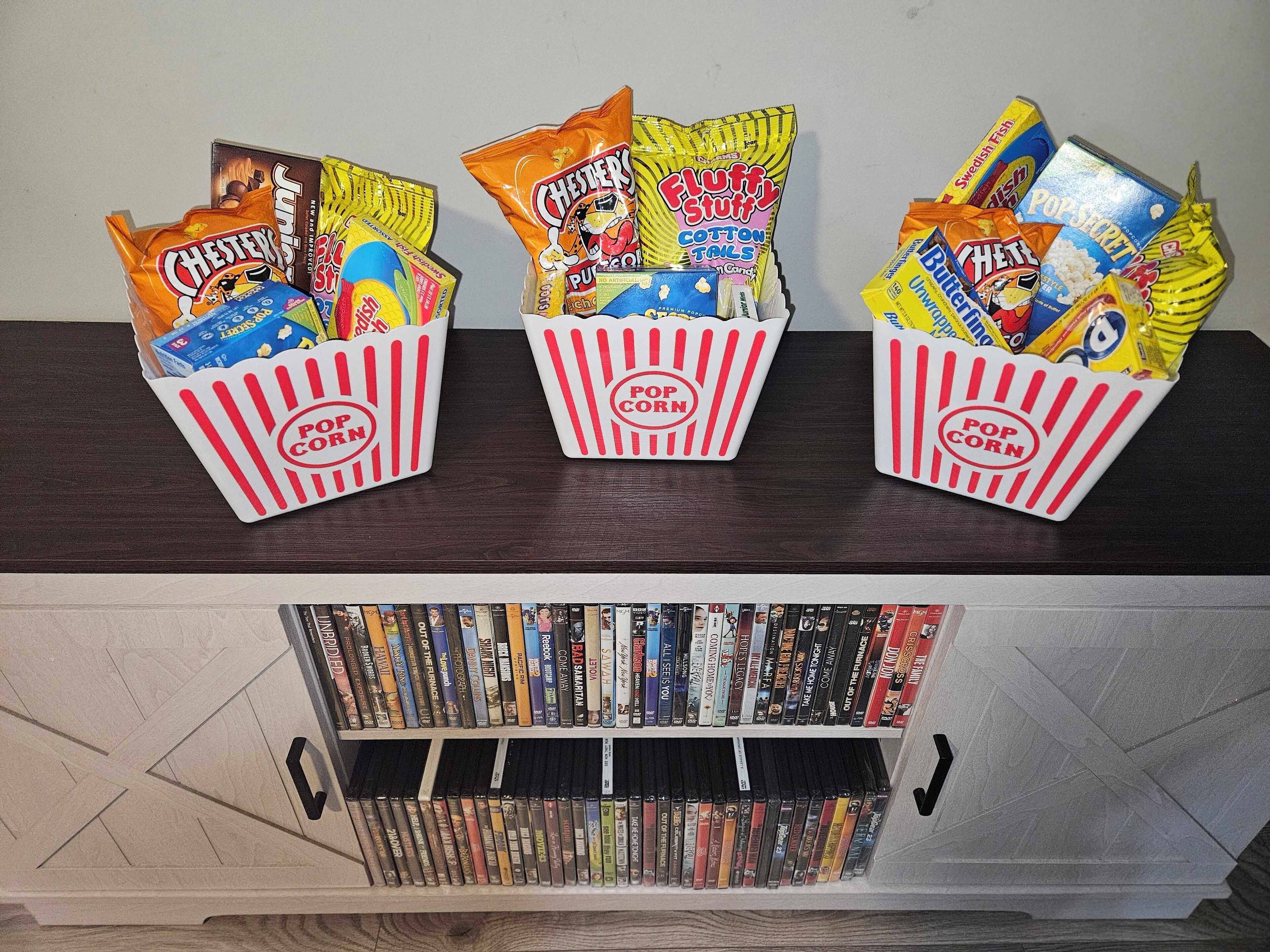 Tres recipientes de palomitas de maíz con diferentes aperitivos sobre un estante de madera. Debajo, un estante para DVD está lleno de numerosos títulos de películas.