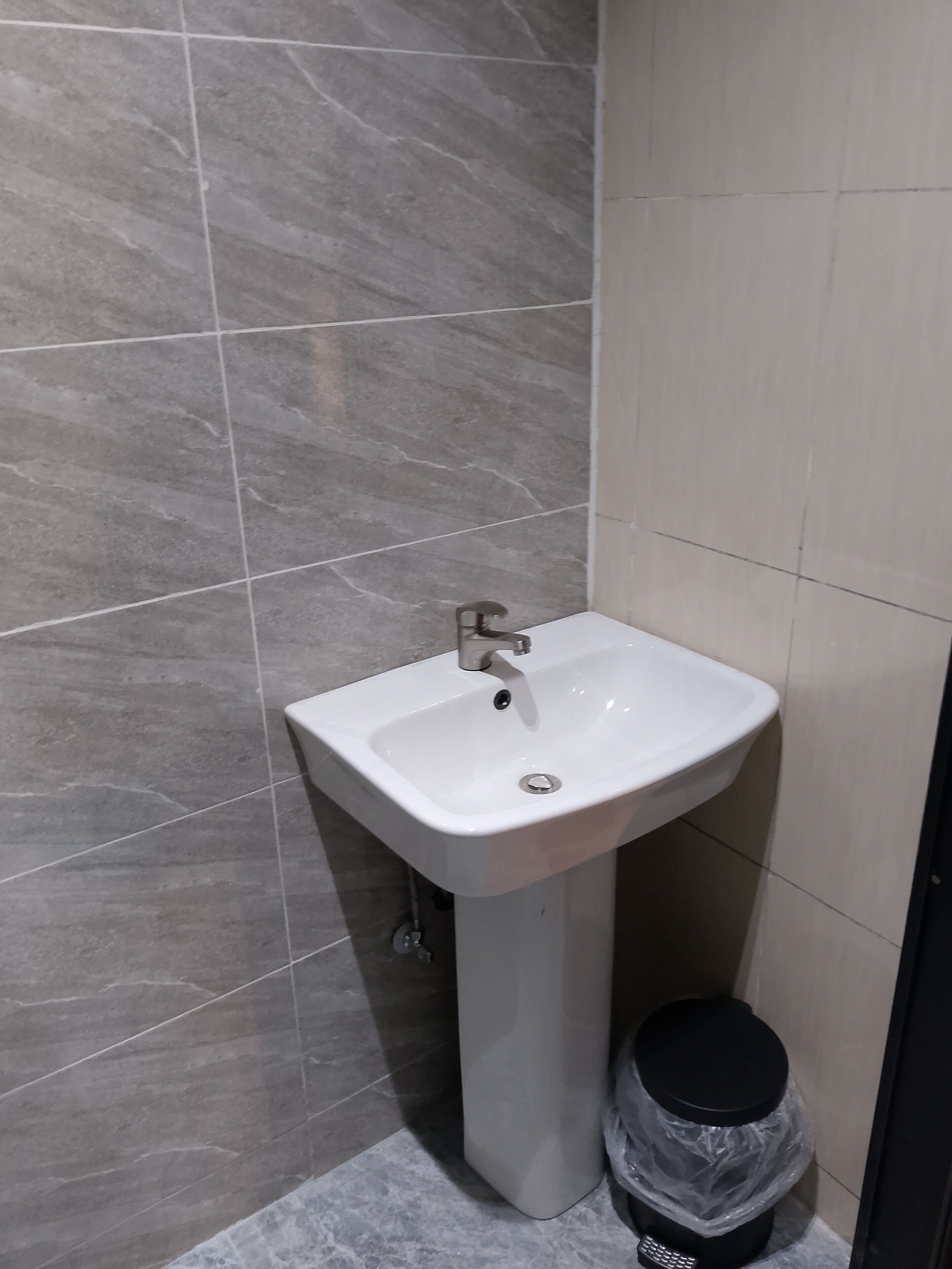 A bathroom with a white pedestal sink, a black trash bin, and grey tiled walls.