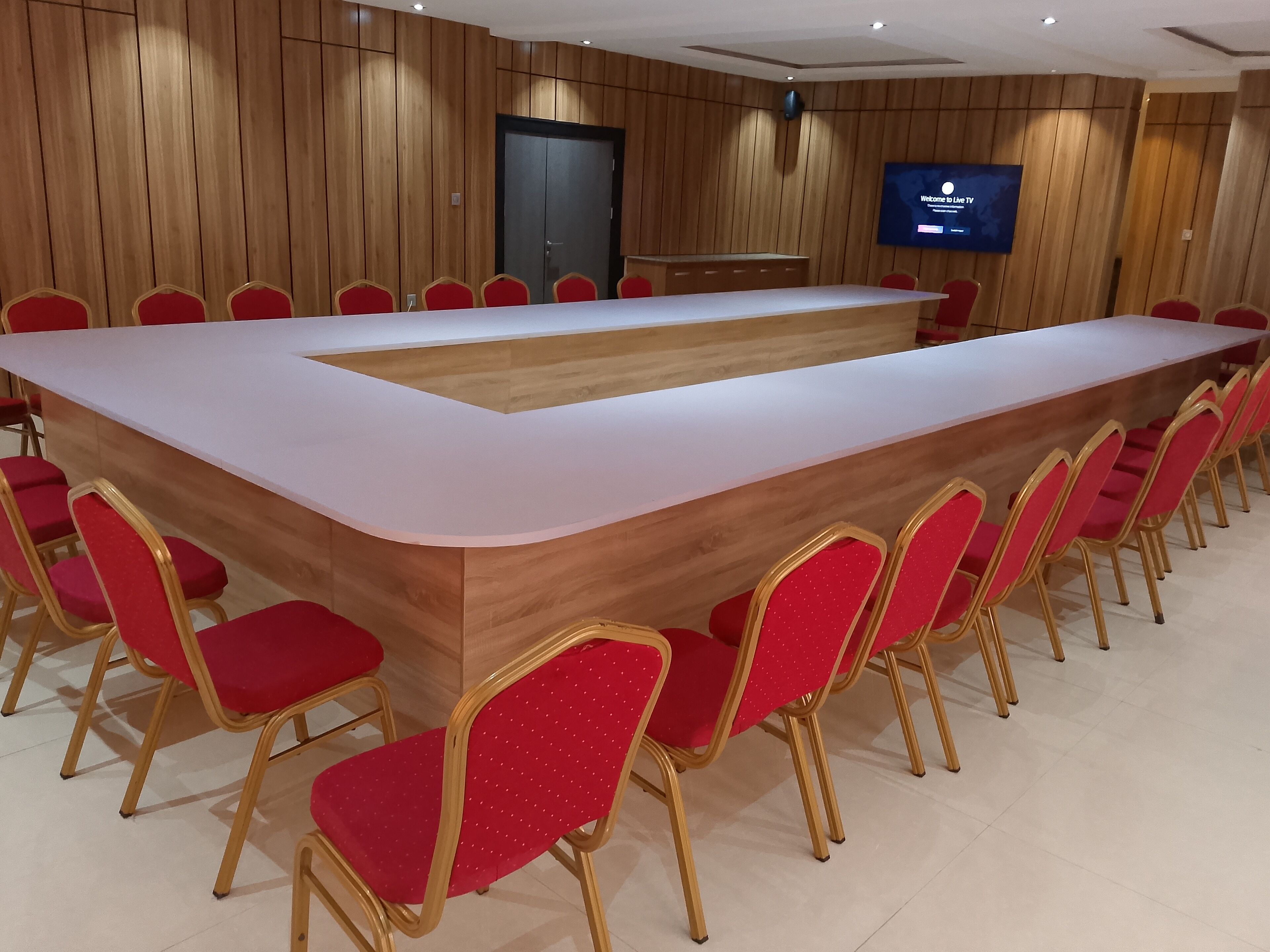 A conference room with a long table and red chairs, a wooden panel wall, and a TV screen displaying "Welcome to Live TV".