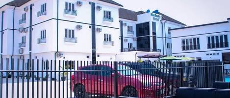 Um hotel de vários andares com varandas, um carro vermelho estacionado na frente e área externa com mesas.