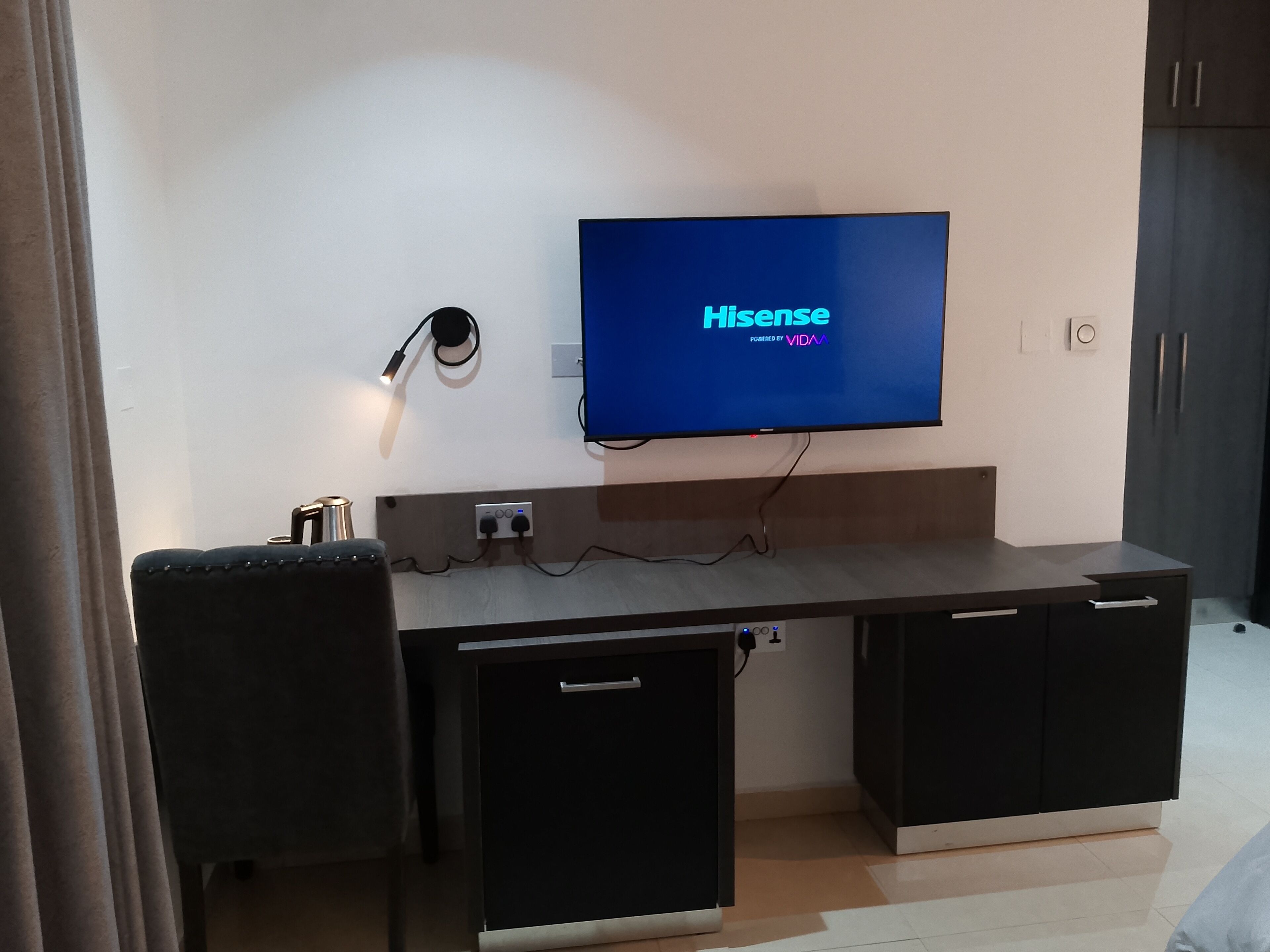 A modern room with a flat-screen TV displaying 'Hisense' and 'Powered by VIVA'. There is a desk with a chair, a lamp, and a cabinet.