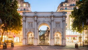 Un portail richement décoré, doté d’une arche centrale et de deux arches latérales, bordé d’arbres et avec des bâtiments en arrière-plan.