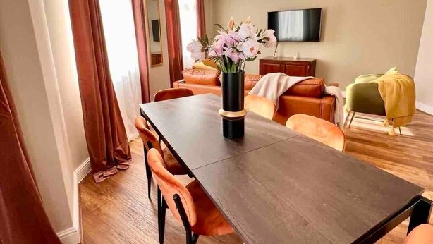 Un salon moderne avec une table à manger en bois foncé, des chaises orange, un canapé et un téléviseur fixé au mur.