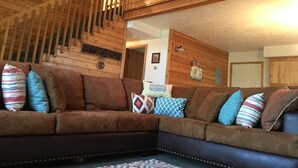 A cozy living room with a brown sectional sofa, wooden walls, and a staircase.